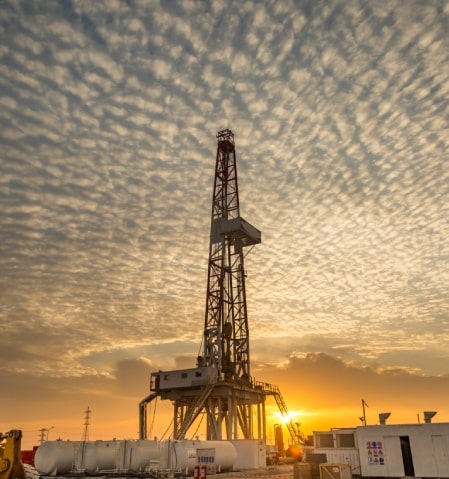 A tall oil drilling rig stands against a dramatic sky filled with dense, low-lying clouds during sunset. The orange hues from the setting sun enhance the striking silhouette of the industrial structure. Tanks and other equipment are visible in the foreground.
